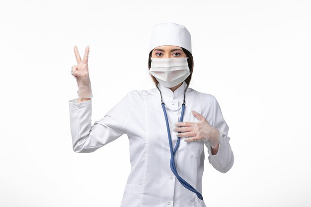 Front view female doctor in white medical suit with a mask due to pandemic on the light wall medicine pandemic virus covid-