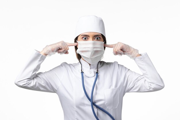 Front view female doctor in white medical suit with a mask due to pandemic shutting her ears on the white desk disease medicine virus pandemic covid-