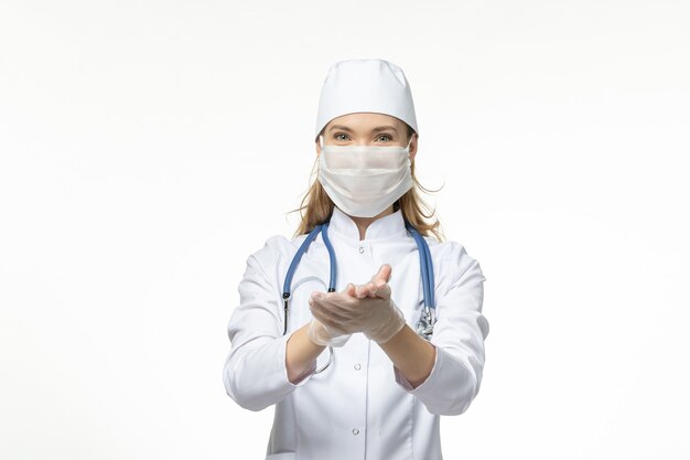 Front view female doctor in white medical suit with protective mask due to coronavirus on light white wall illness covid- pandemic disease