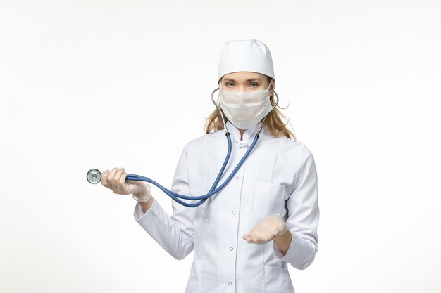 Front view female doctor in white medical suit with sterile mask due to coronavirus using a stethoscope on white desk illness covid- pandemic disease