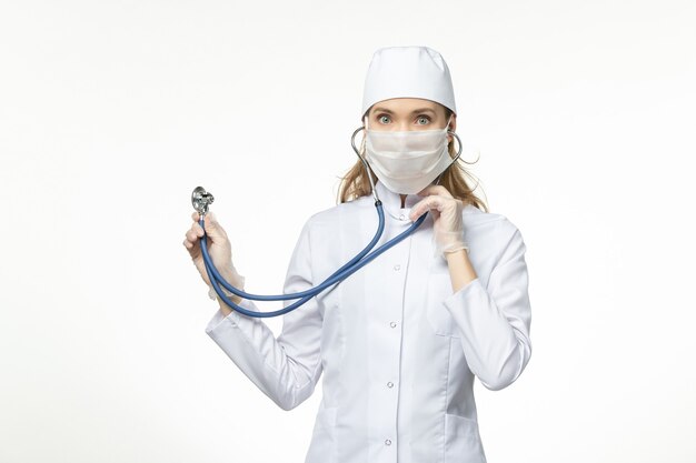 Вид спереди женщина-врач в белом медицинском костюме со стерильной маской из-за коронавируса с помощью стетоскопа на белой стене болезнь пандемическая болезнь covid-