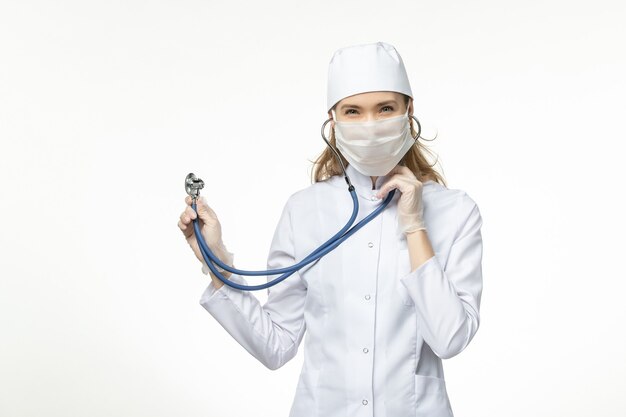 Front view female doctor in white medical suit with sterile mask due to coronavirus using a stethoscope on white wall illness pandemic disease covid-