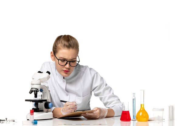 Front view female doctor in white medical suit working with microscope on light white space