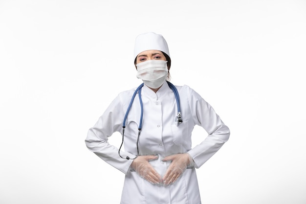 Free photo front view female doctor in white sterile medical suit and wearing mask due to covid- having stomachache on white wall illness virus covid- pandemic disease