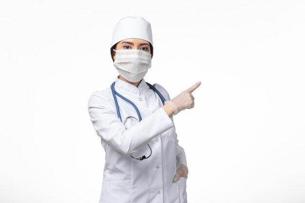 Front view female doctor in white sterile medical suit and wearing mask due to covid- posing on light white wall illness virus covid- pandemic disease