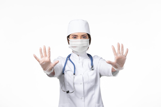 Front view female doctor in white sterile medical suit and wearing mask due to covid- posing on the white wall illness virus covid- pandemic disease