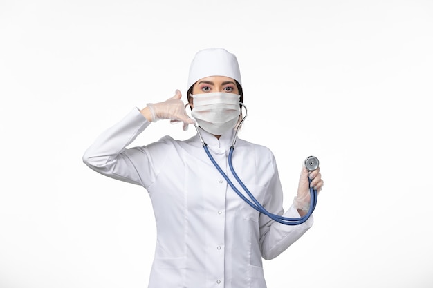 Front view female doctor in white sterile medical suit and wearing mask due to covid- using stethoscope posing on white wall illness virus covid- pandemic disease