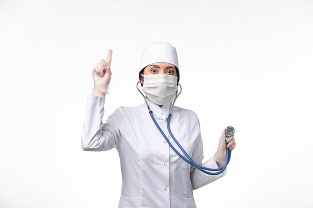 흰색 멸균 의료 복을 입은 여성 의사와 covid- 흰색 책상에 청진기를 사용하여 마스크 착용 전면보기 질병 바이러스 covid- pandemic disease