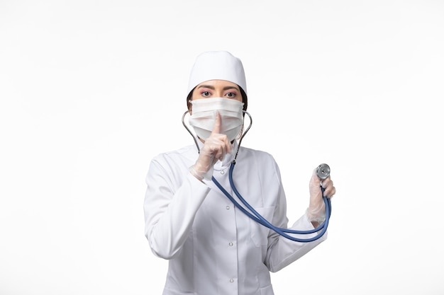 Front view female doctor in white sterile medical suit and wearing mask due to covid- using stethoscope on the white wall illness covid- pandemic disease