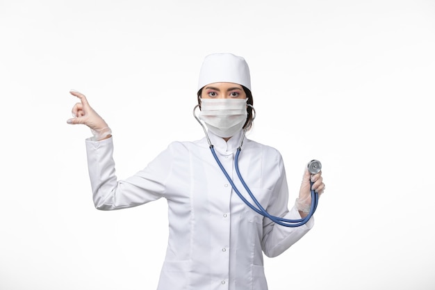 Вид спереди женщина-врач в белом стерильном медицинском костюме и в маске из-за covid - с помощью стетоскопа на белой стене, вируса болезни, covid - пандемического заболевания