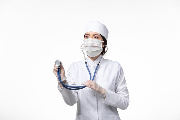 흰 벽에 청진기를 사용하여 흰색 멸균 의료 복을 입고 마스크를 착용 한 전면보기 여성 의사
