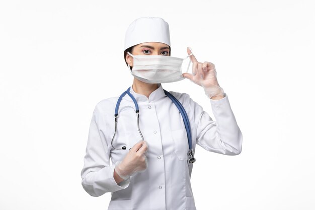 Вид спереди женщина-врач в белом стерильном медицинском костюме в маске из-за заболевания коронавирусом на белой стене.