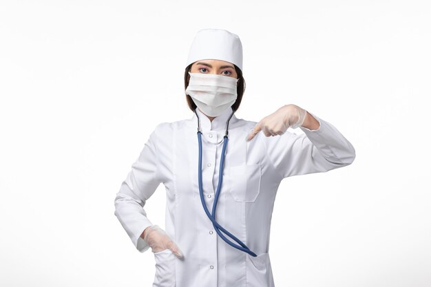 Front view female doctor in white sterile medical suit with mask due to coronavirus on the light white wall disease illness pandemic virus covid-