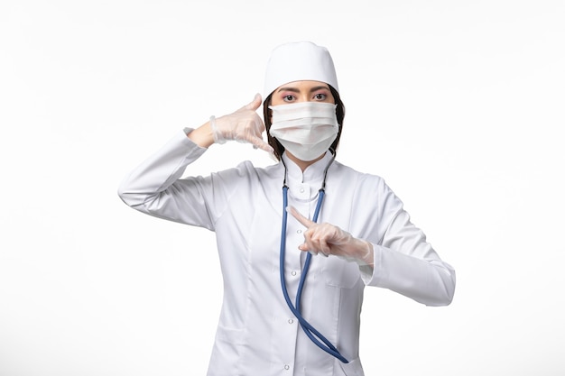 Front view female doctor in white sterile medical suit with mask due to coronavirus on light-white wall illness pandemic disease covid- virus