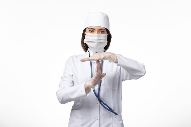 Front view female doctor in white sterile medical suit with mask due to coronavirus posing on white wall illness pandemic disease covid- virus