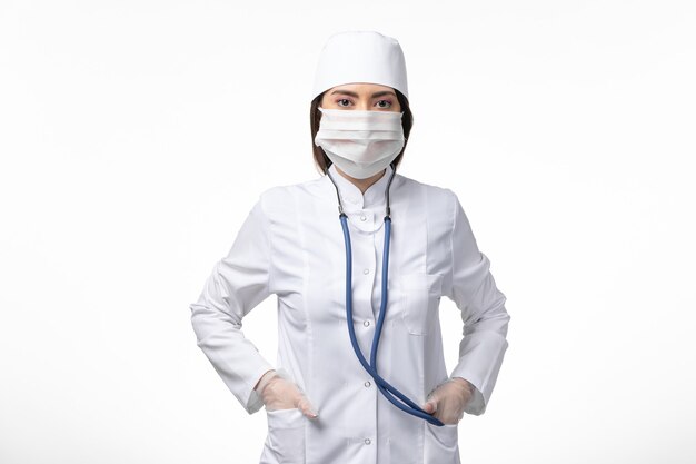 Front view female doctor in white sterile medical suit with mask due to coronavirus on white wall illness pandemic disease covid- virus