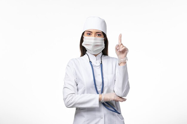Вид спереди женщина-врач в белом стерильном медицинском костюме с маской из-за коронавируса, подняв палец на белой стене, болезнь, болезнь, пандемический вирус