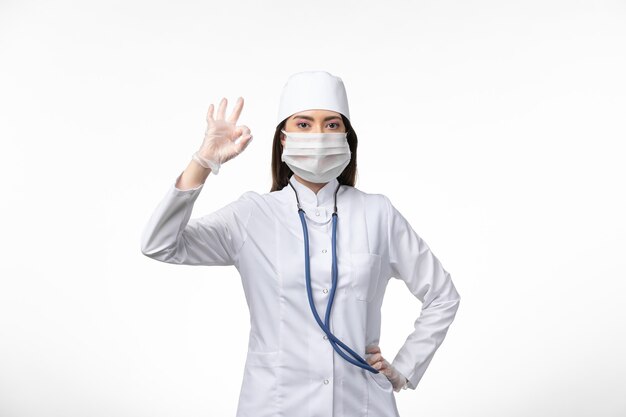 Foto gratuita medico femminile di vista frontale in vestito medico sterile bianco con maschera a causa della malattia da virus pandemico della malattia del muro bianco