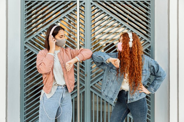 Front view of female friends with face masks outdoors doing the elbow salute