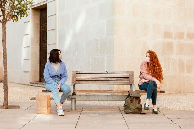 Front view of female friends with face masks outdoors sitting on a bench