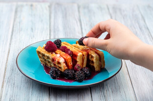 Front view of female hand over sweet waffles with blackberry on a blue plate on a gray surface