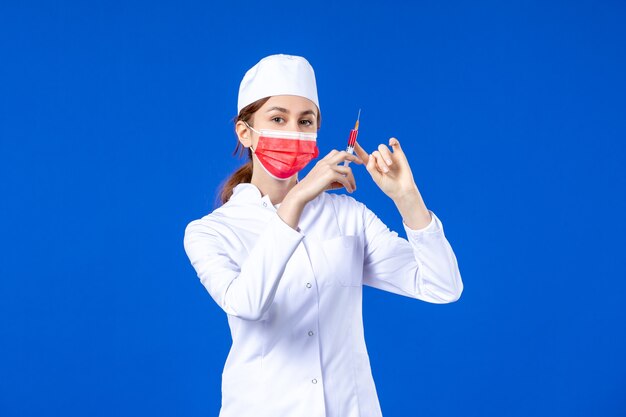 Front view female nurse in white medical suit with red mask and injection in her hands on blue 