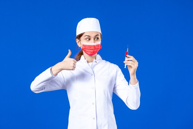 Front view female nurse in white medical suit with red mask and injection in her hands on blue 