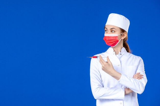 Front view female nurse in white medical suit with red mask and injection in her hands on blue 