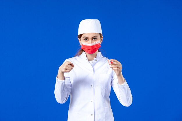 Front view female nurse in white medical suit with red mask and injection in her hands on blue 