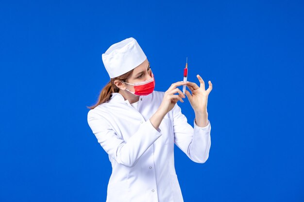 Front view female nurse in white medical suit with red mask and injection in her hands on blue 