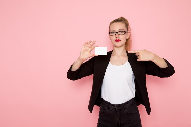 Front view of female office worker in black strict jacket holding card on light-pink wall