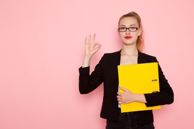 Front view of female office worker in black strict jacket holding documents on the light pink wall