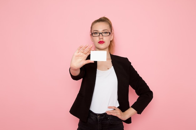 Front view of female office worker in black strict jacket holding plastic card on light-pink wall