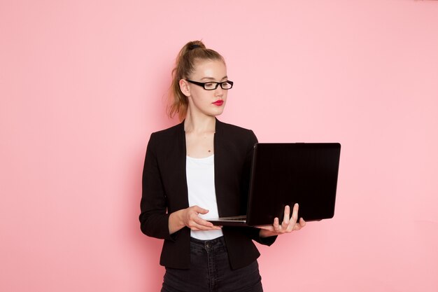 Front view of female office worker in black strict jacket holding and using her laptop on light-pink wall