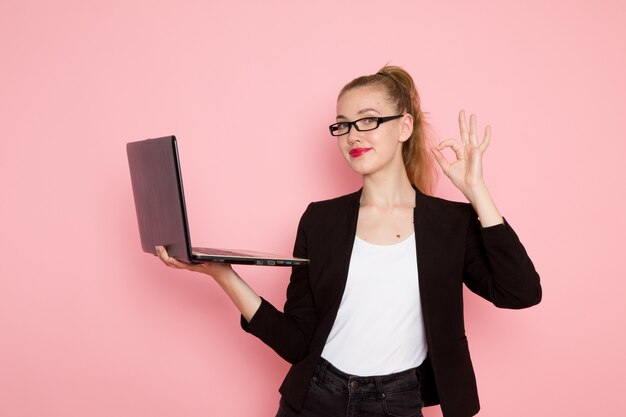 Front view of female office worker in black strict jacket holding and using laptop on the light-pink wall