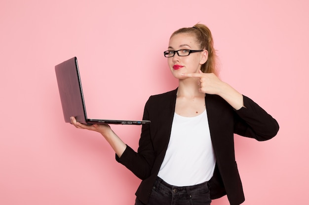 Front view of female office worker in black strict jacket holding using laptop on light-pink wall