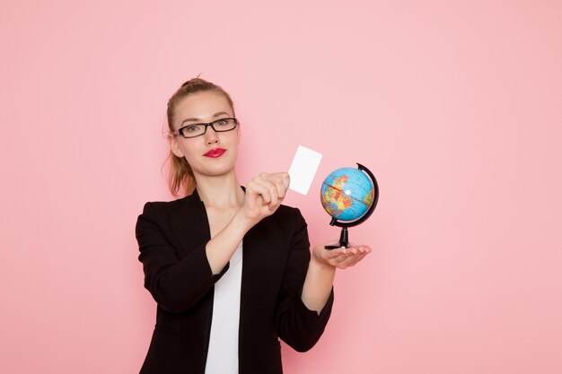 Вид спереди офисного работника в черной строгой куртке, держащего белую карточку и глобус на светло-розовой стене