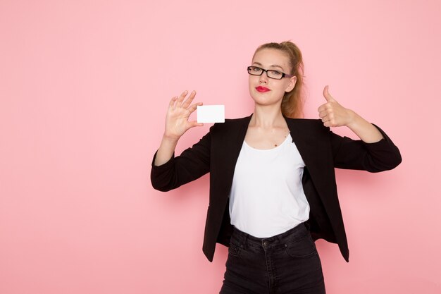 Front view of female office worker in black strict jacket holding white card smiling on light-pink wall