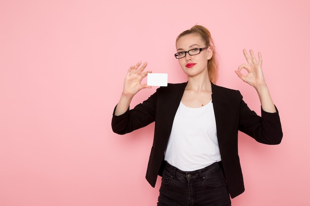 Front view of female office worker in black strict jacket holding white plastic card on light-pink wall