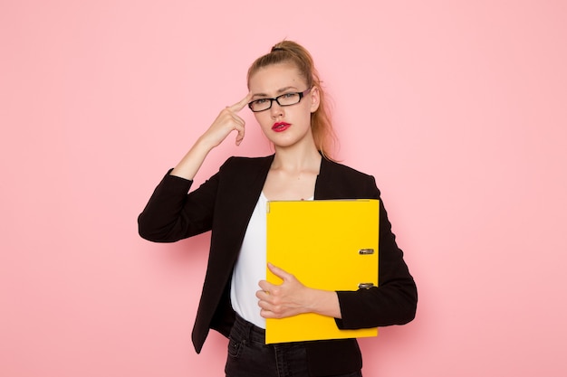 Front view of female office worker in black strict jacket holding yellow document on the pink wall