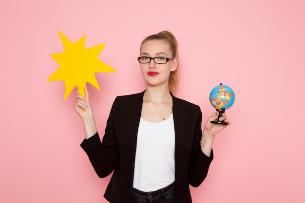 Vista frontale dell'impiegato femminile in giacca rigorosa nera che tiene cartello giallo e globo sulla parete rosa