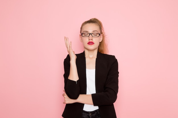 Front view of female office worker in black strict jacket posing and closing her eyes on light-pink wall