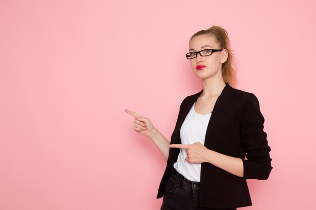 Front view of female office worker in black strict jacket posing pointing out on light-pink wall