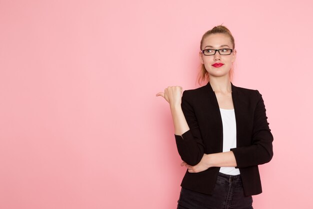 Vista frontale dell'impiegato femminile in giacca rigorosa nera in posa e sorridente sulla parete rosa chiaro