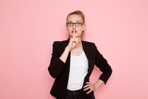 Front view of female office worker in black strict jacket showing silence sign on pink wall