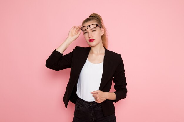 Front view of female office worker in black strict jacket taking off her sunglasses on pink wall