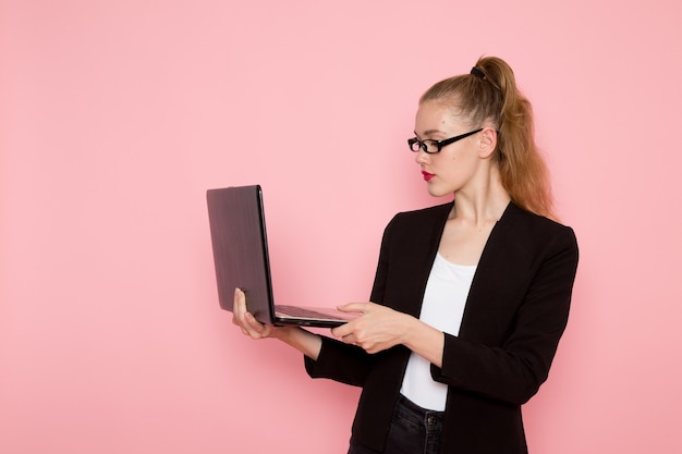 Front view of female office worker in black strict jacket using her laptop on pink wall