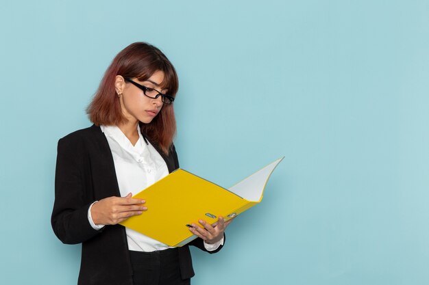 Front view female office worker holding and reading yellow file on blue surface