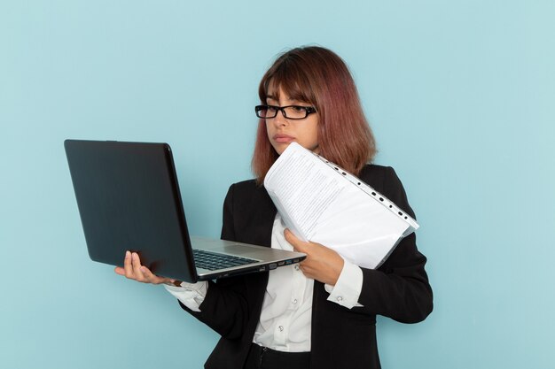 Front view female office worker in strict suit holding documents and laptop on blue surface