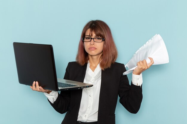 Front view female office worker in strict suit holding papers and laptop on blue surface